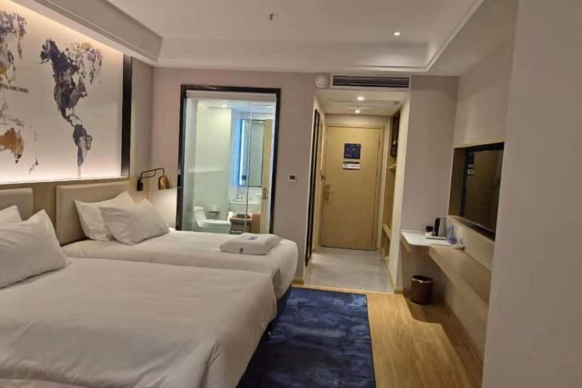 A hotel room with two beds, a world map on the wall, a TV, and a bathroom visible through a glass door.