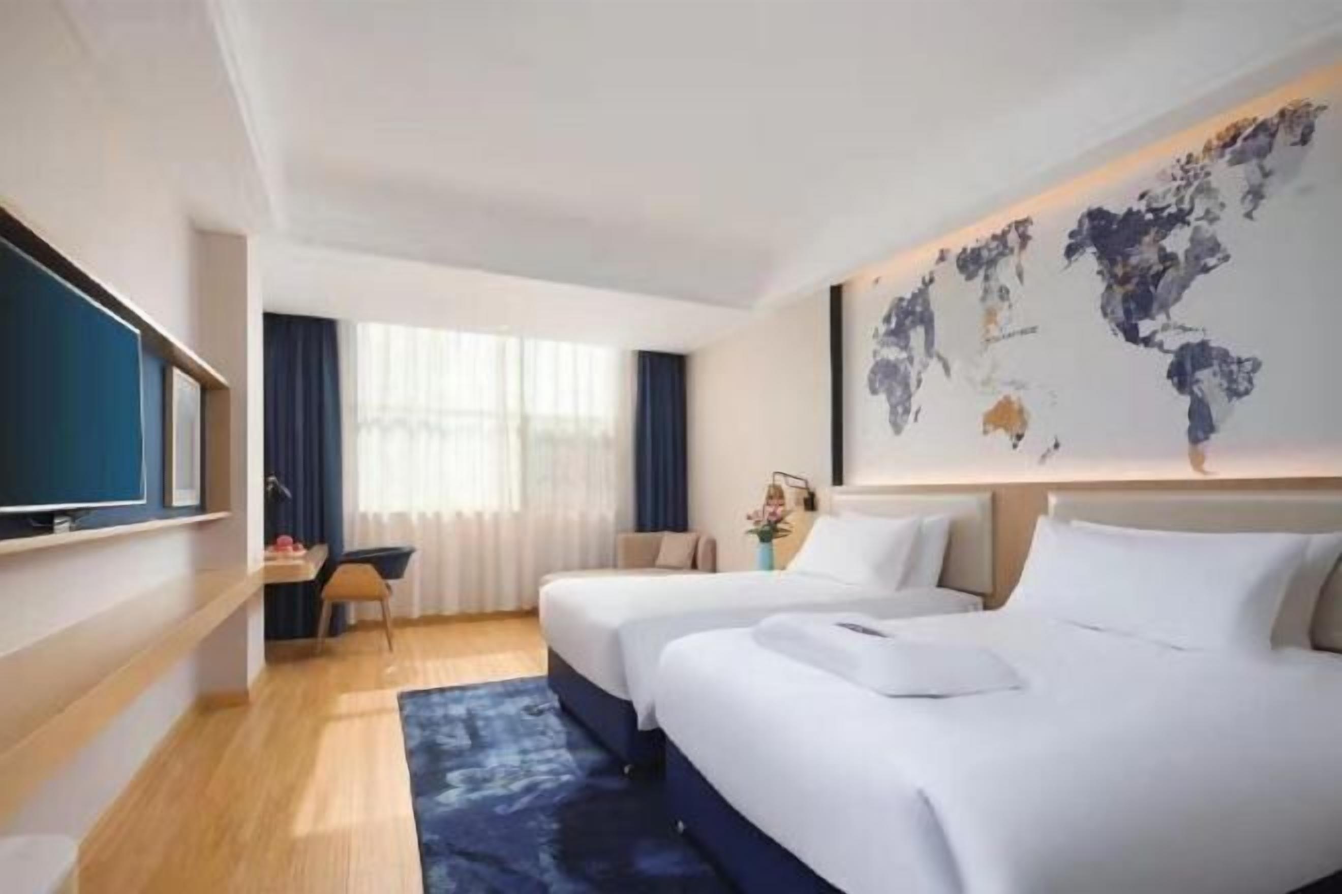 Une chambre d’hôtel avec deux lits, une grande carte du monde accrochée au mur, un bureau avec une chaise et un téléviseur placé à gauche.