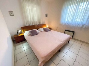 Ein Schlafzimmer mit einem Bett, zwei Nachttischen, einem Tischchen, einem Fenster mit Vorhängen und einer Heizung.