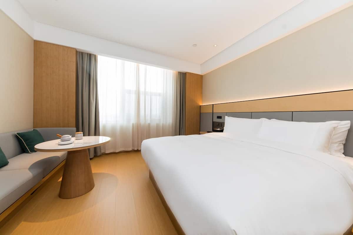A modern hotel room with a large bed, a round table with a chair, a sofa, and a large window with curtains.