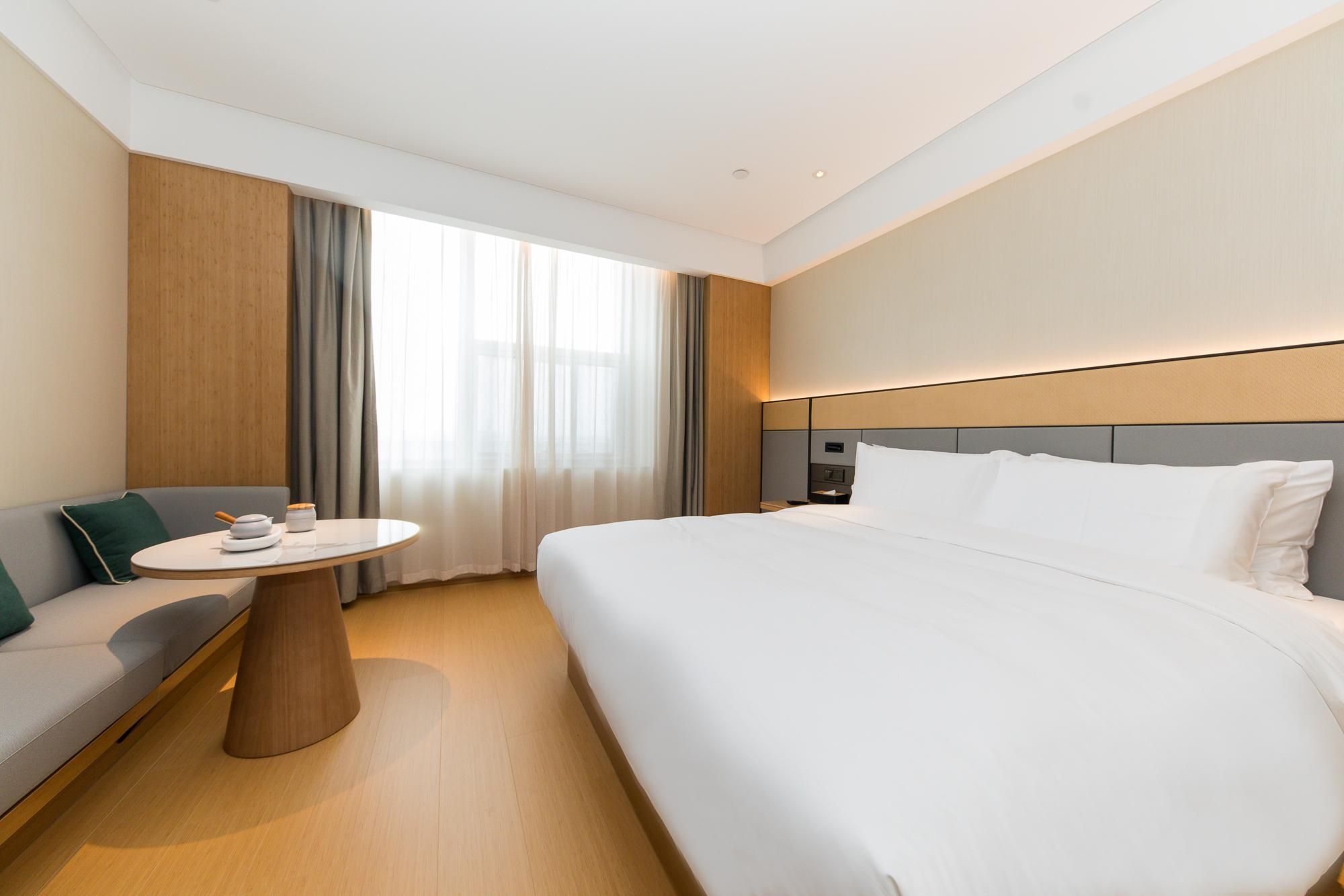 Une chambre d’hôtel moderne avec un grand lit, une table ronde avec une chaise, un canapé et une grande fenêtre avec des rideaux.