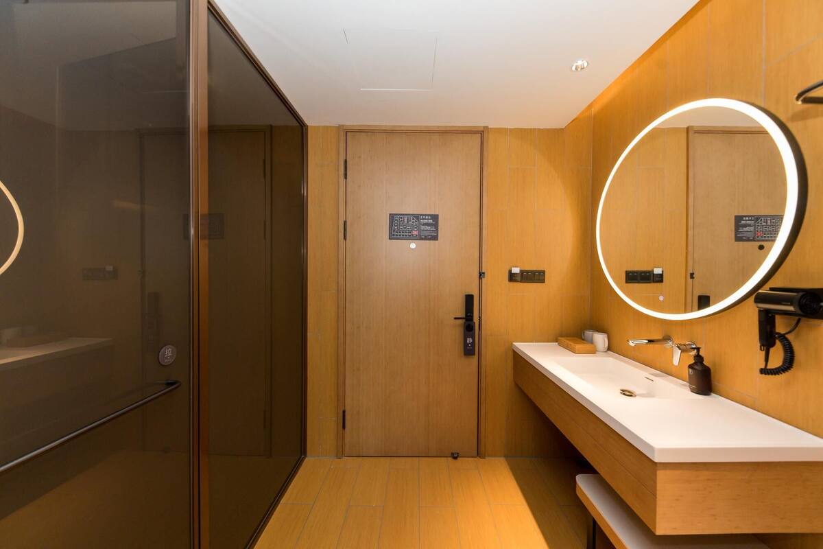A modern bathroom with a wooden vanity, a mirror with a circular light, and a glass shower enclosure.