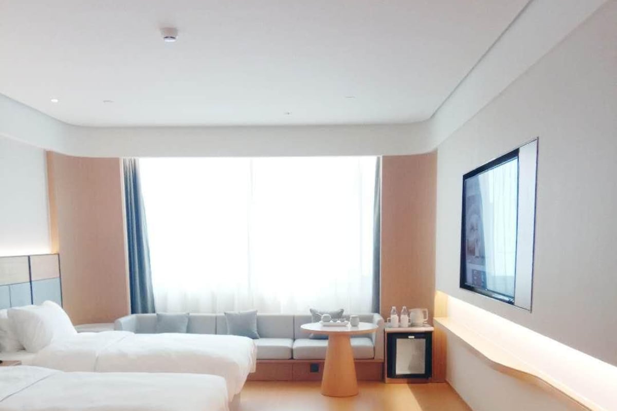 A hotel room with two beds, a small table, a TV on the wall, and a window with curtains.