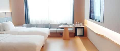 A hotel room with two beds, a small table, a TV mounted on the wall, and a window with curtains.