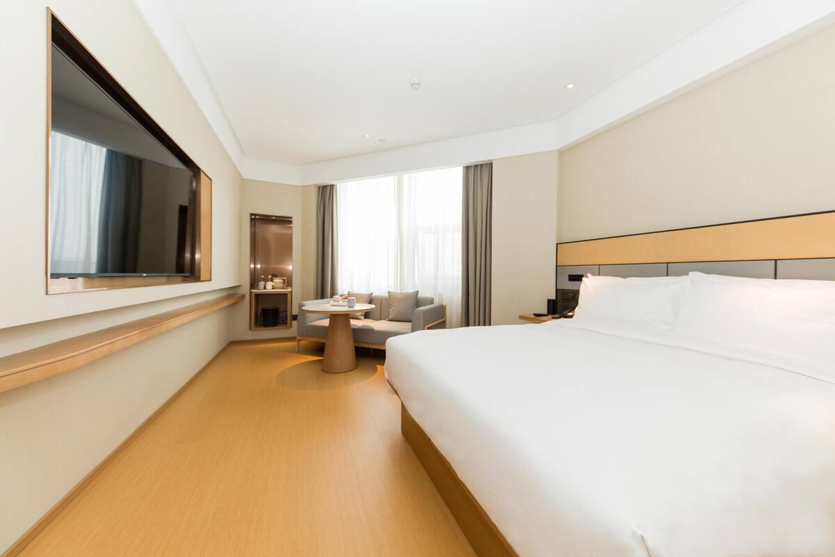 A modern hotel room with a large bed, a small round table, a sofa, and a television mounted on the wall.