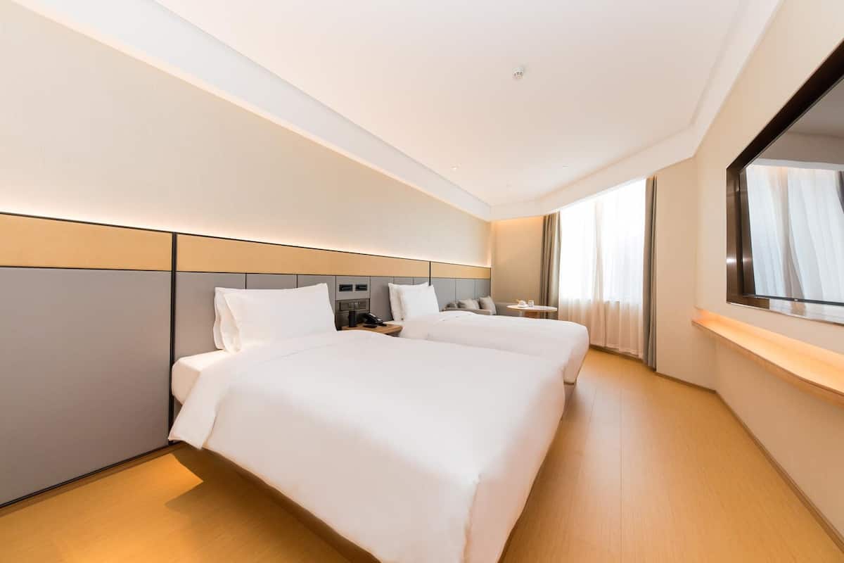 A modern hotel room with two beds, a large mirror, and a view of the city.