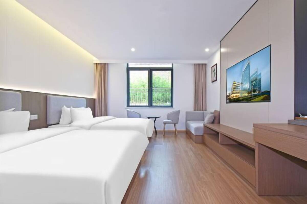 A hotel room with two beds, a desk, a chair, a small table, and a large window with a view of greenery.