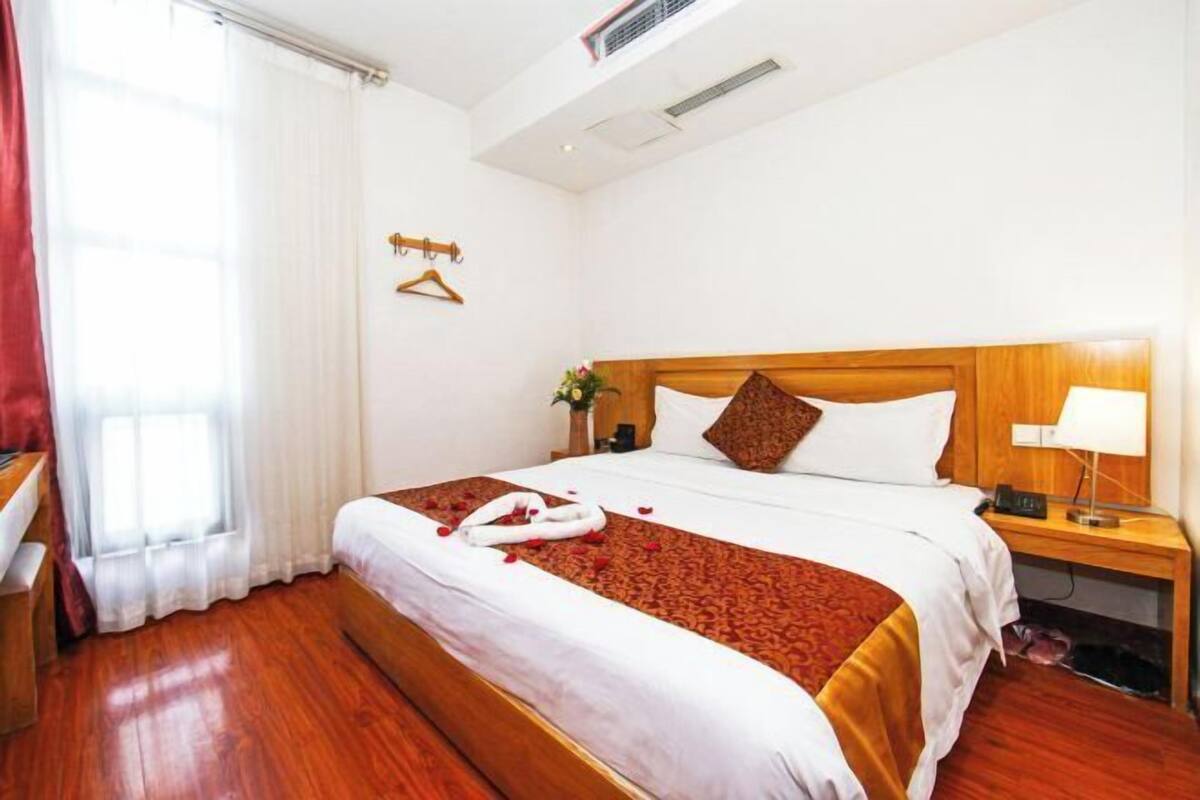A hotel room with a wooden bed, a bedside table with a lamp, a hanging coat rack, and a window with curtains.