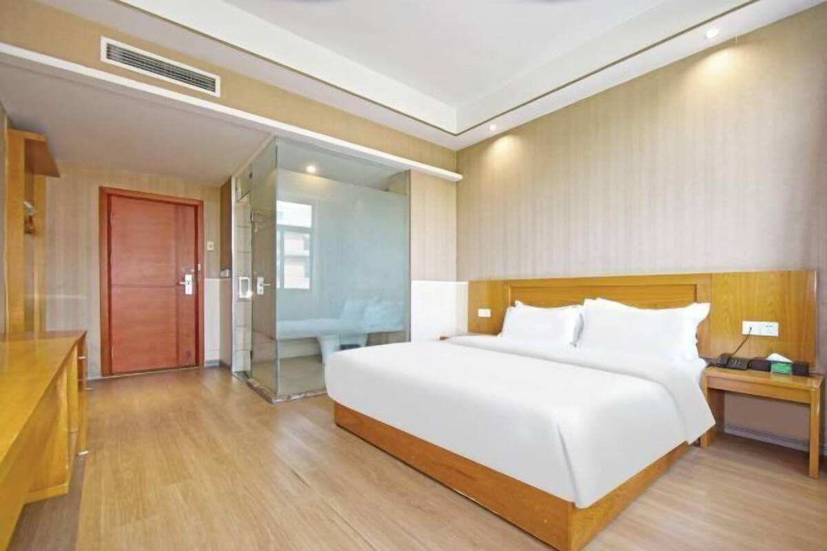 A hotel room with a wooden bed, a bedside table, a nightstand, a wooden bench, and a glass door leading to a bathroom.