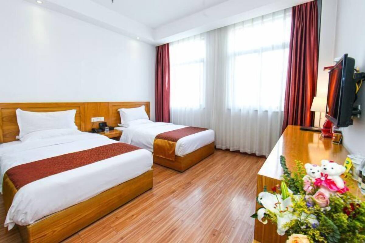 A hotel room with two beds, a wooden headboard, a desk with a lamp, and a window with curtains.