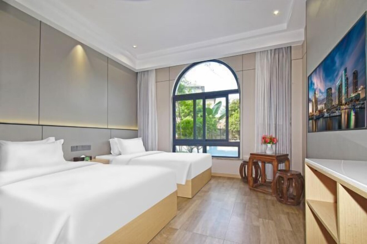 A hotel room with two beds, a large window with a view of greenery, a painting on the wall, and a small table with flowers.