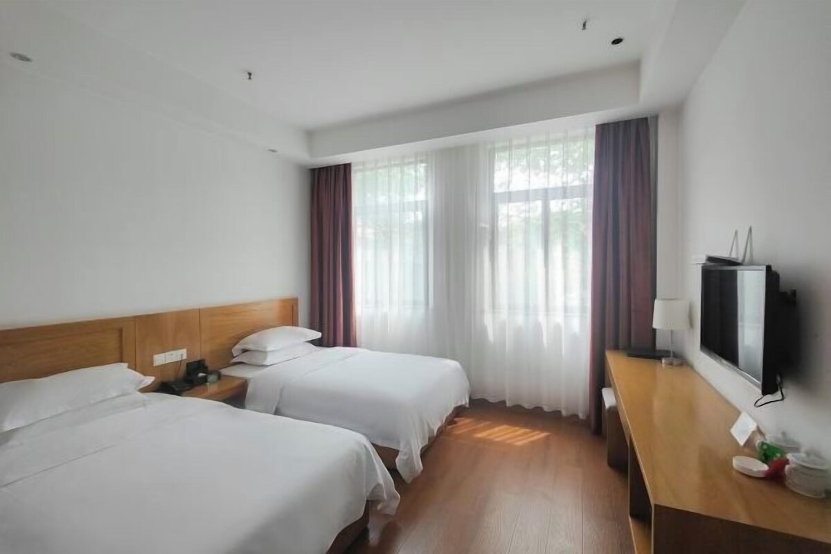 A hotel room with two beds, a wooden headboard, a desk with a TV, and a window with sheer curtains.
