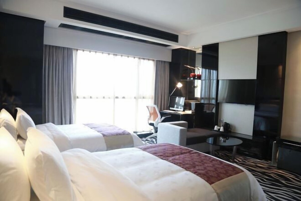 A hotel room with two beds, a desk, a chair, a TV, and a large window with curtains.