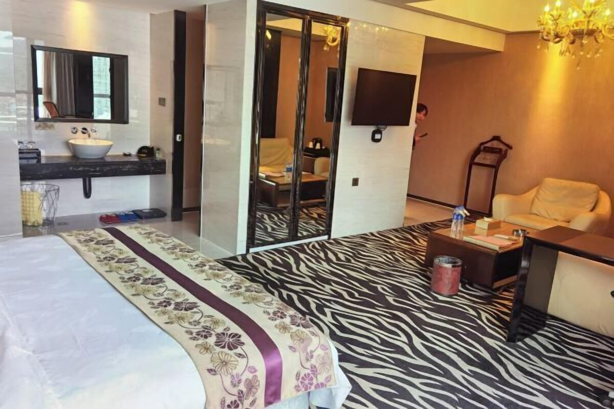 A hotel room with a bed, a bathroom with a sink and mirror, a television, a sofa, and a patterned carpet.