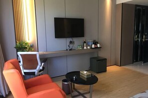 A modern hotel room with a flat-screen TV mounted on the wall, a small table with a bottle and glass, a chair, and a trash can. - Mellow Crystal Hotel (Zhangjiajie)