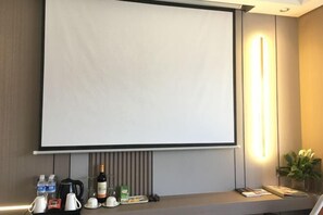A hotel room with a large screen for presentations, a coffee station with various beverages, and a bedside table with a plant and a book. - Mellow Crystal Hotel (Zhangjiajie)
