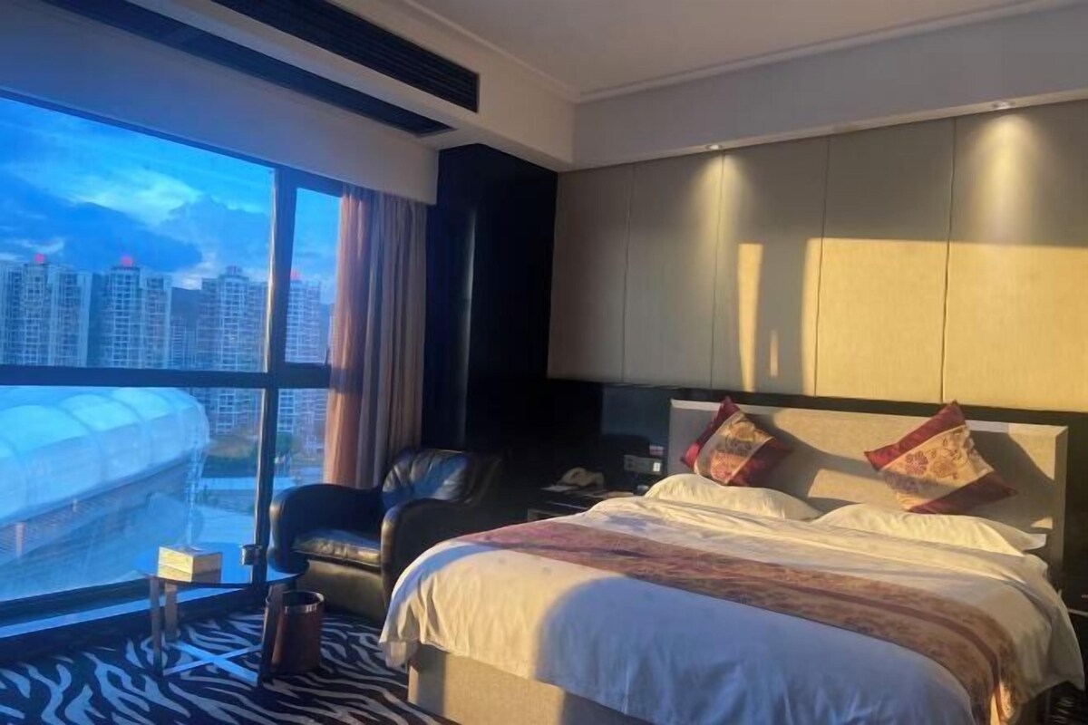 A hotel room with a large bed, a chair, a desk, and a view of a cityscape through large windows.
