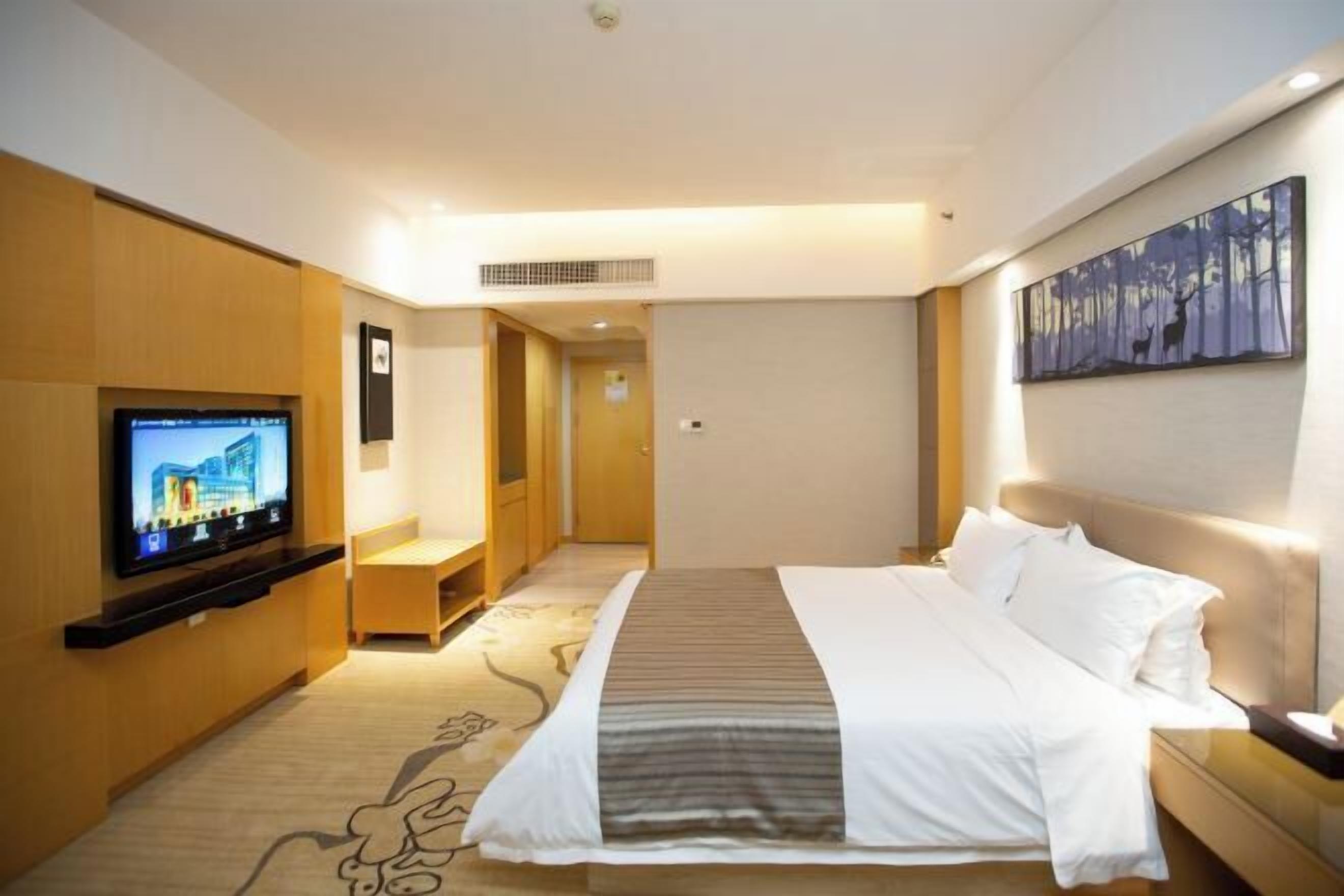 A hotel room with a bed, a TV mounted on the wall, a painting on the wall, and a carpet with a pattern.