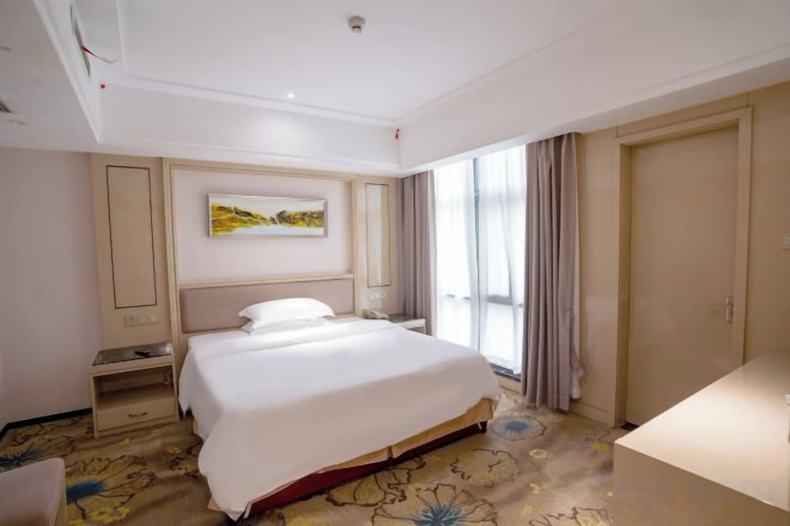 A hotel room with a large bed, a bedside table, a nightstand, a window with curtains, and a door.