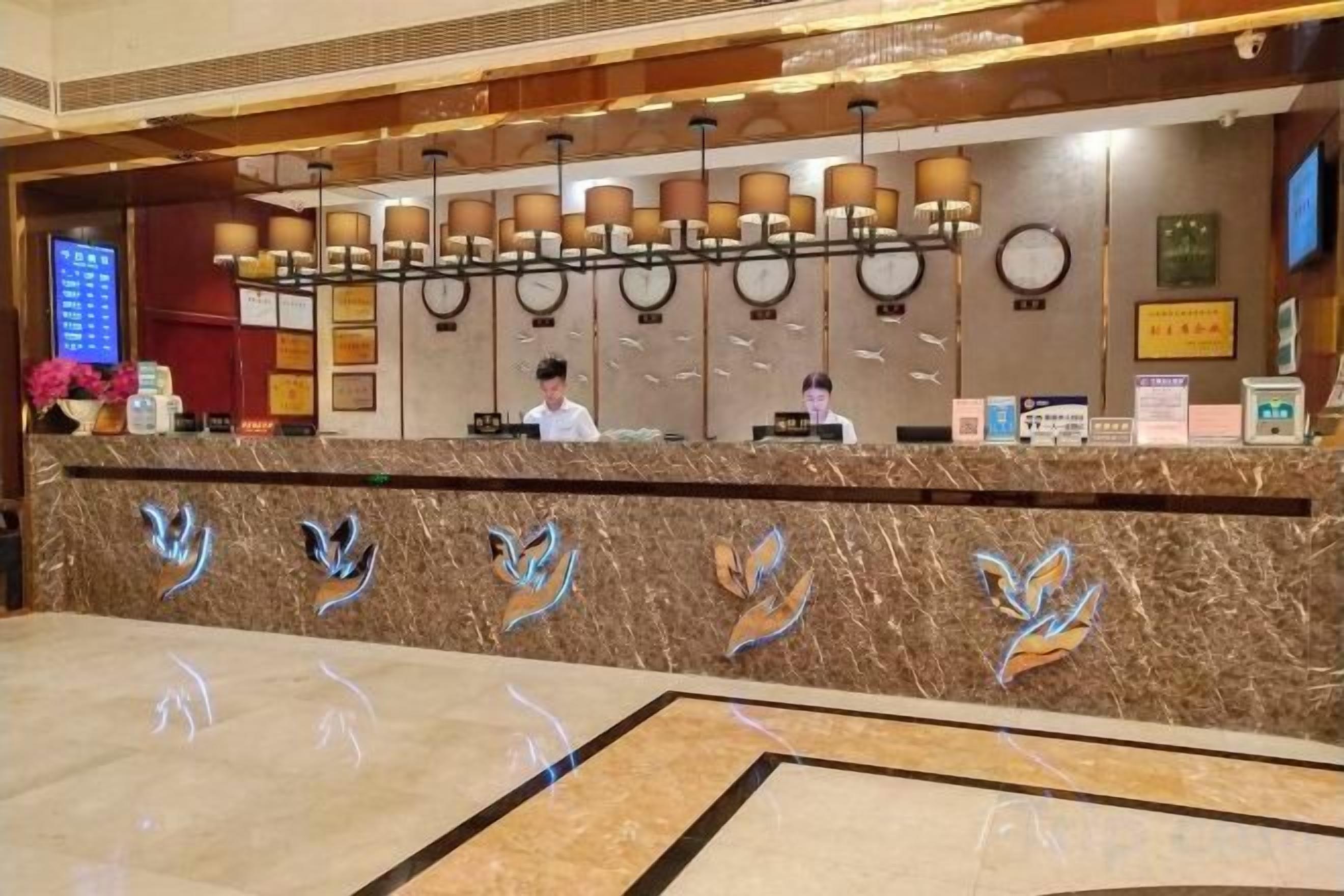 A hotel reception area with a marble counter, a digital screen, and a row of lamps.