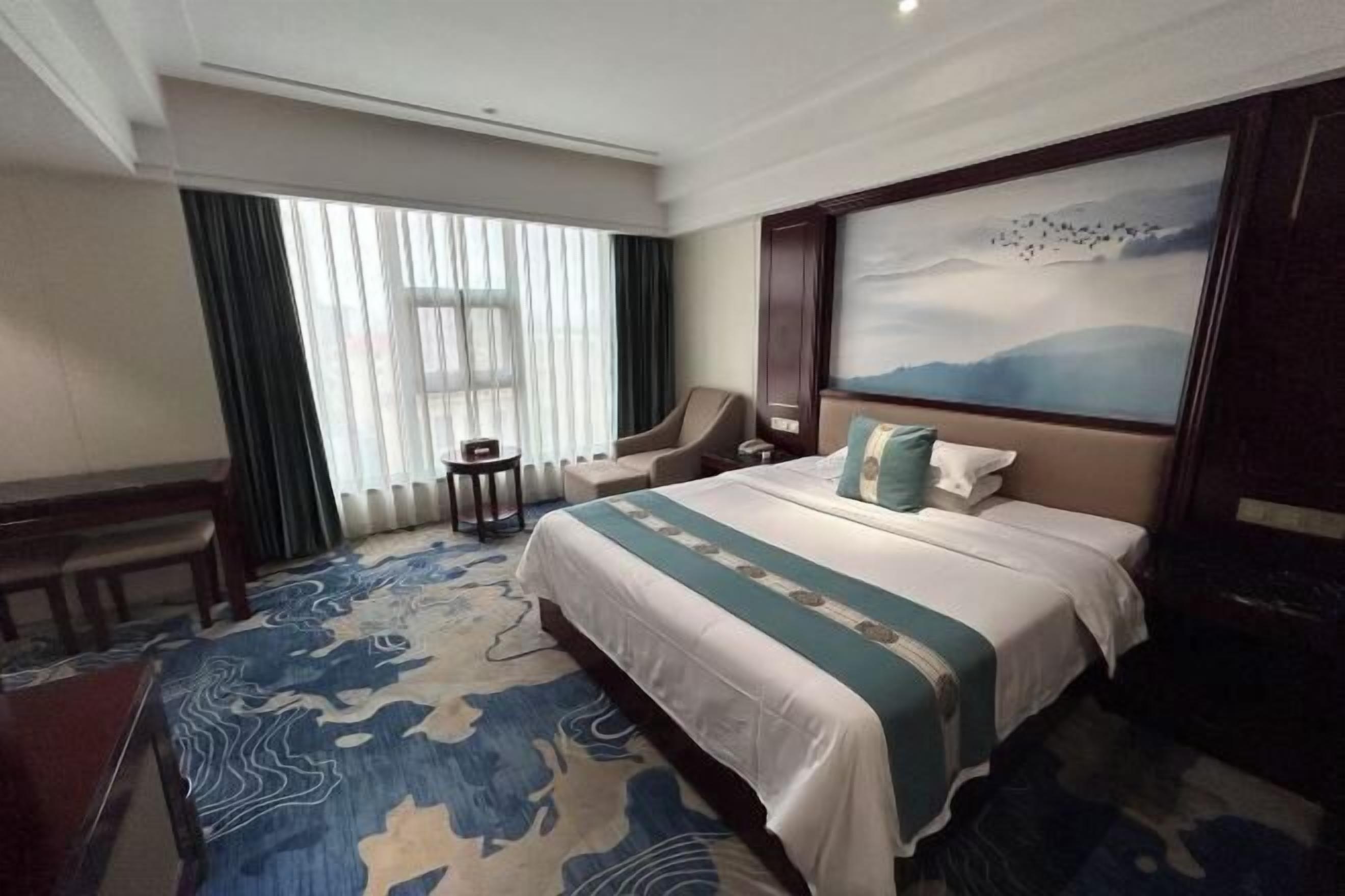 A hotel room with a large bed, a chair, a small table, and a painting on the wall.