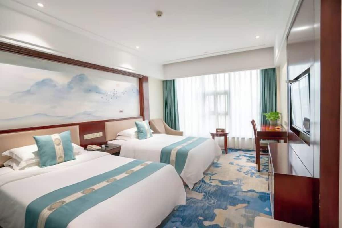 Quarto de hotel com duas camas, uma pintura grande na parede, uma escrivaninha com cadeira e uma televisão.