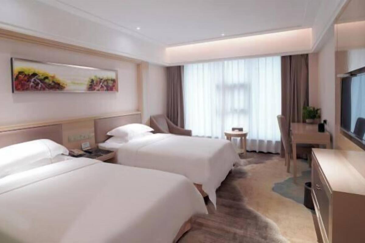 Quarto de hotel com duas camas, uma escrivaninha, uma cadeira, televisão e uma janela grande com cortinas.