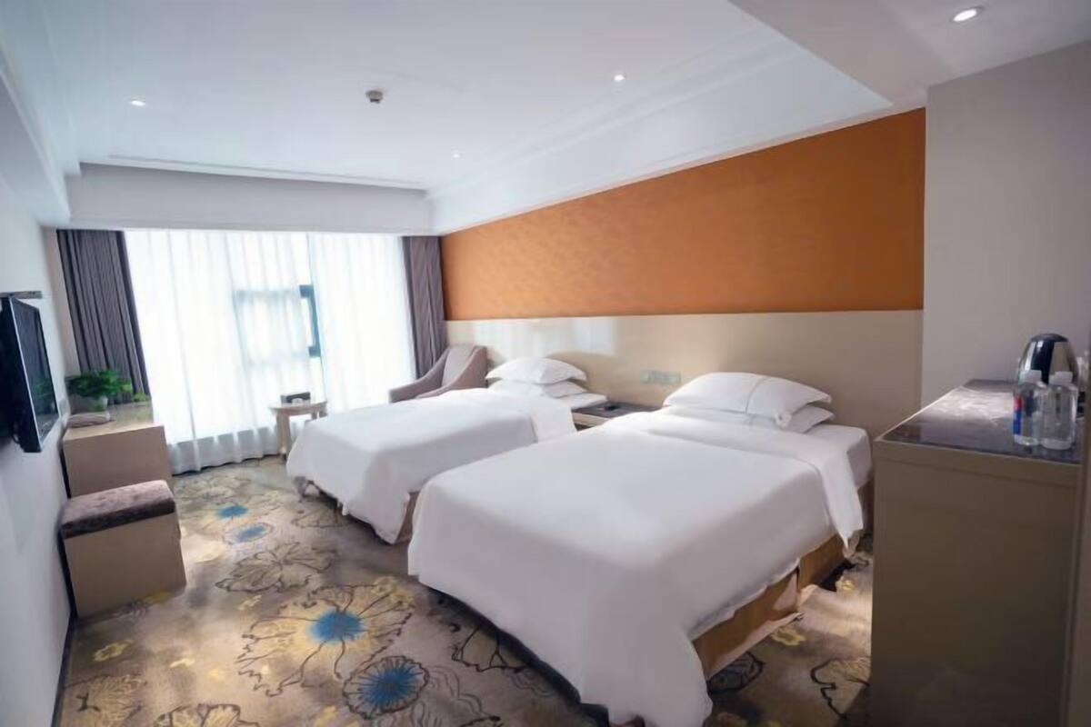 Quarto de hotel com duas camas, uma escrivaninha, uma cadeira, uma televisão e uma janela grande com cortinas.