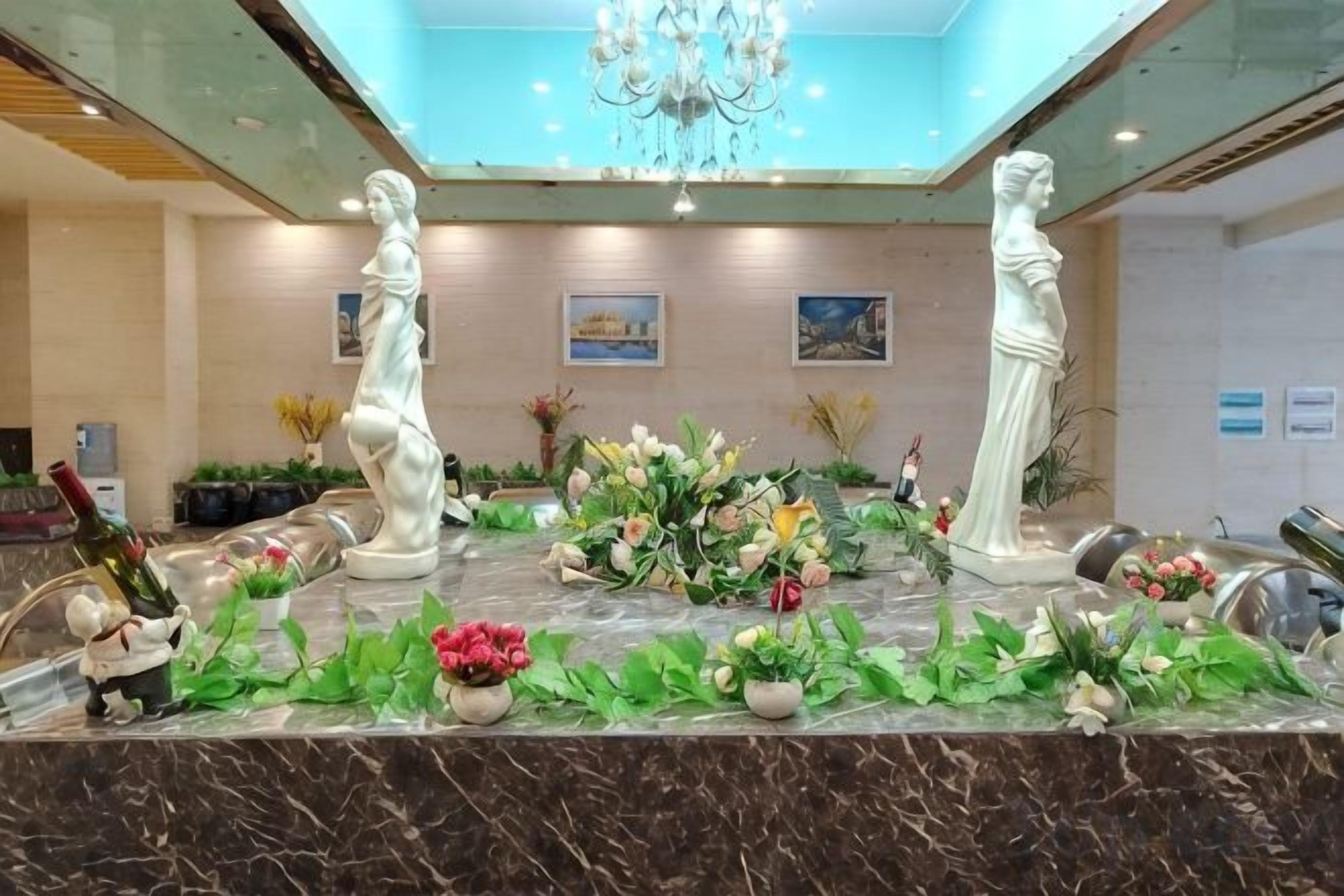 A marble reception desk with two white statues, a chandelier, and floral arrangements.
