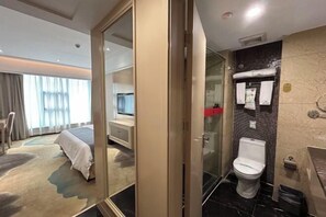 Habitación de hotel con cama, baño con inodoro, ducha y cabina de ducha con paredes de vidrio.