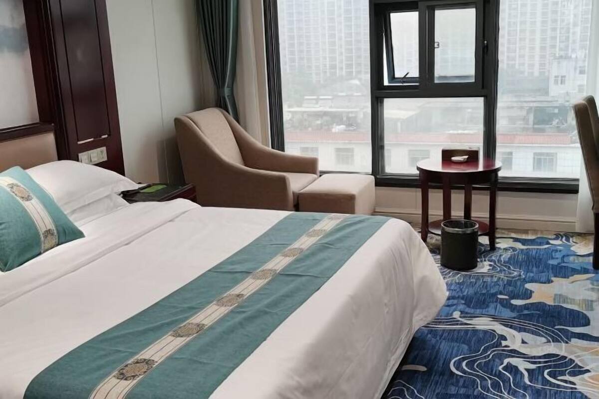 Quarto de hotel com cama, uma cadeira, uma mesinha e janela com vista para edifícios.