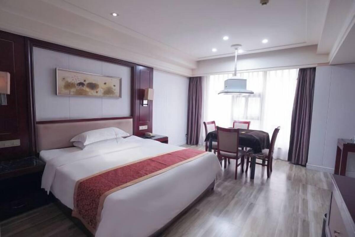 Quarto de hotel com uma cama grande, uma escrivaninha com cadeira, uma janela com cortinas e um quadro na parede.