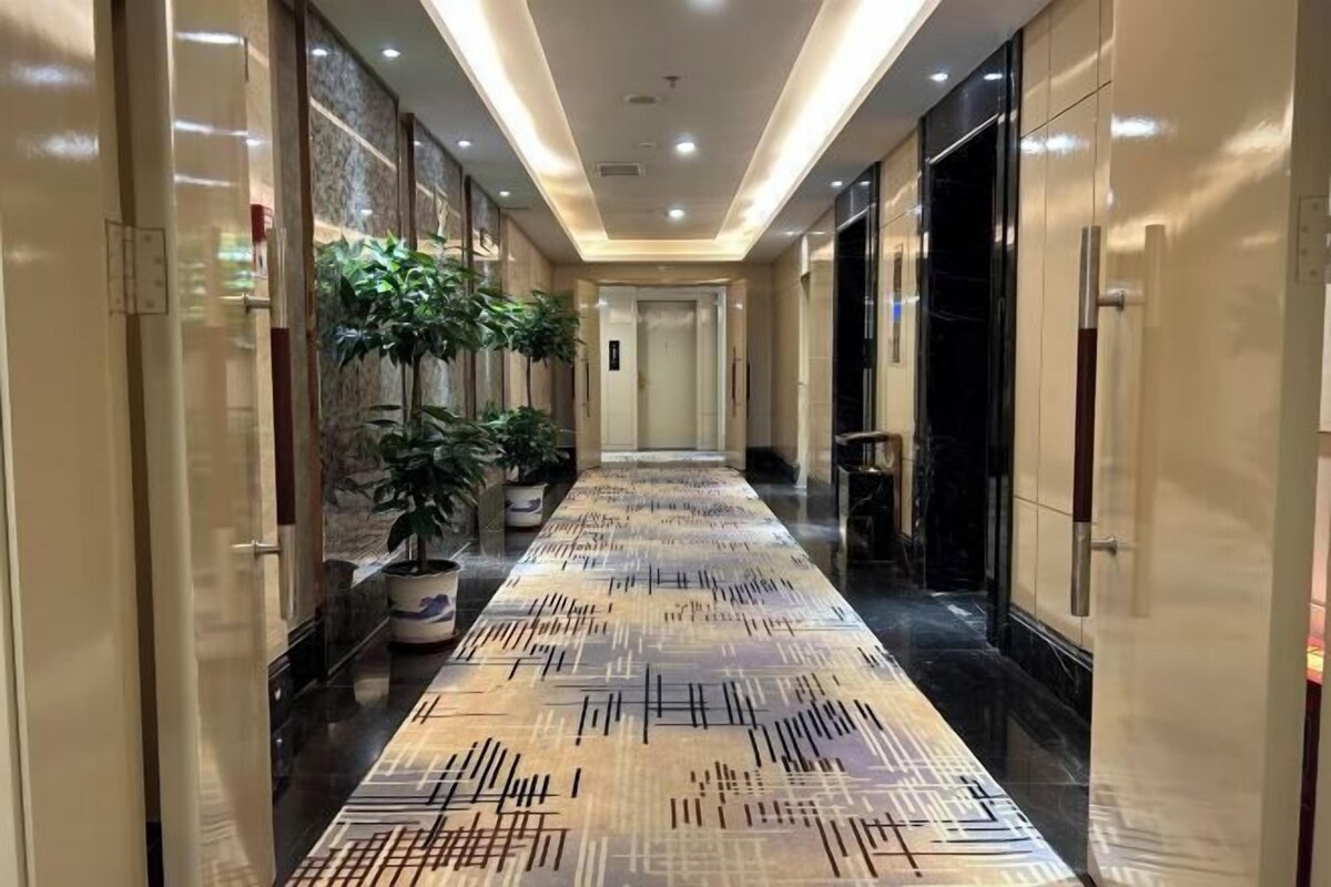 Pasillo de hotel con alfombra estampada, plantas en macetas y un ascensor moderno.