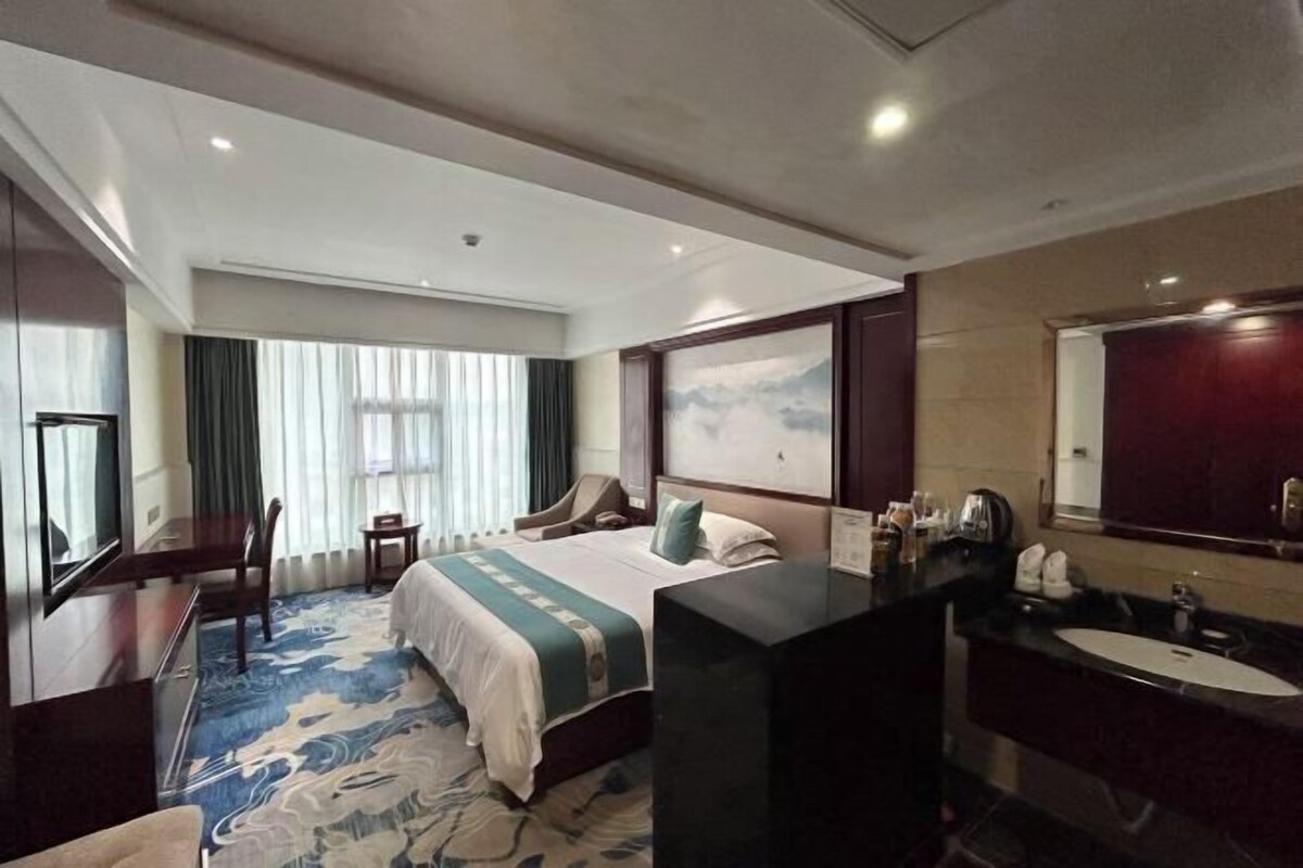 Habitación de hotel con una cama grande, un escritorio, una silla, un televisor, un baño con lavabo y espejo, y una ventana con cortinas.