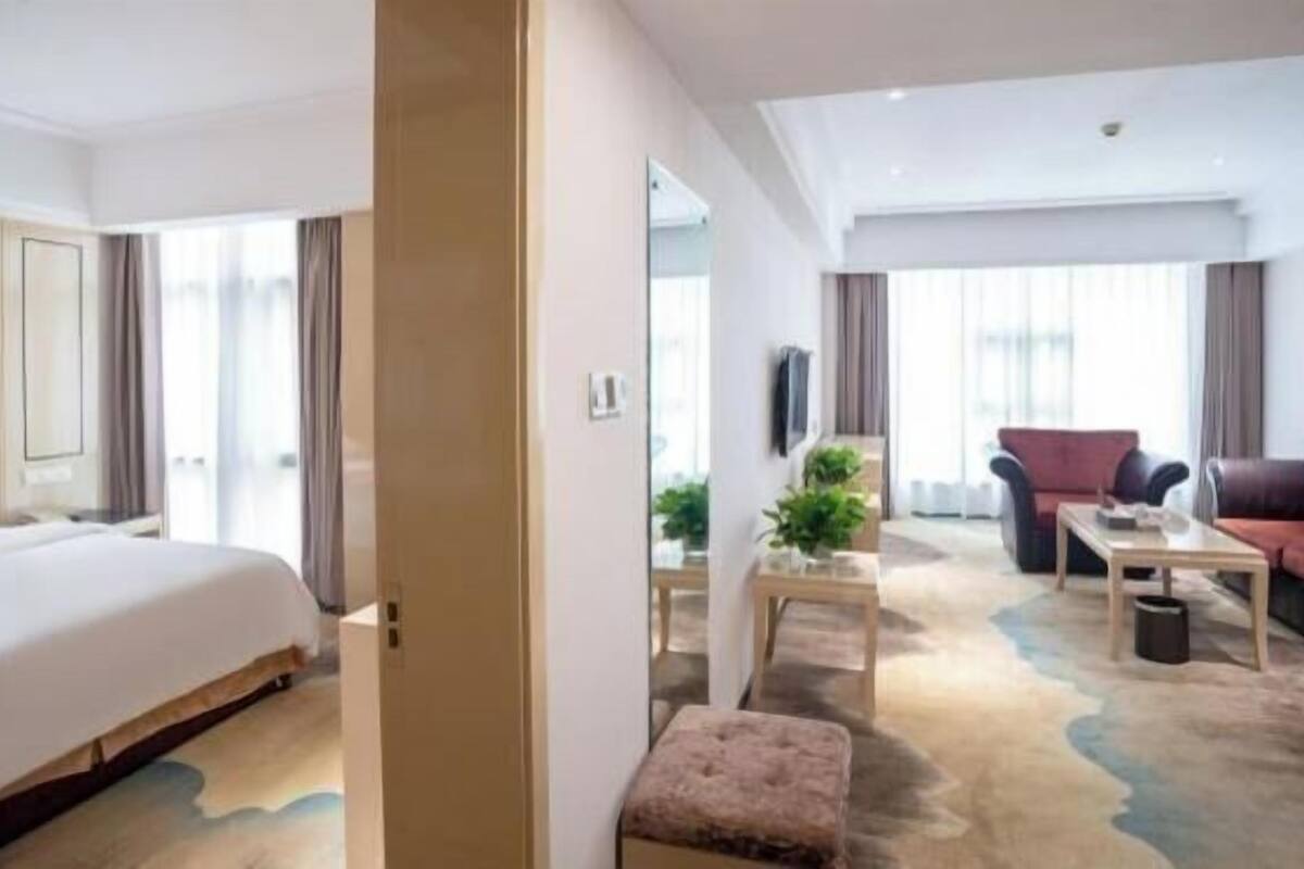 Quarto de hotel com cama, área de estar com sofá e poltronas, mesa pequena e planta em vaso.