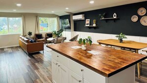 Una cocina moderna con encimera de madera, armarios blancos y una pared de acento negra. Hay plantas, una mesa de comedor y un sofá en la zona de estar contigua.