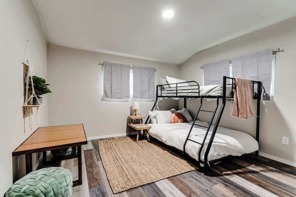 A single-bed room with a bunk bed, a small wooden table, a green ottoman, a woven rug, and a plant on a shelf.