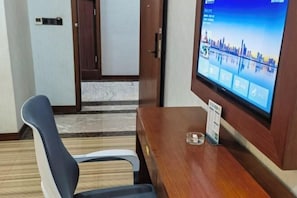 Quarto de hotel com uma escrivaninha de madeira, uma cadeira, televisão exibindo uma vista urbana e uma porta que dá para outro quarto.