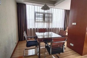 Quarto de hotel com piso de madeira, uma janela grande com cortinas, uma mesa de jantar com cadeiras e um lustre.