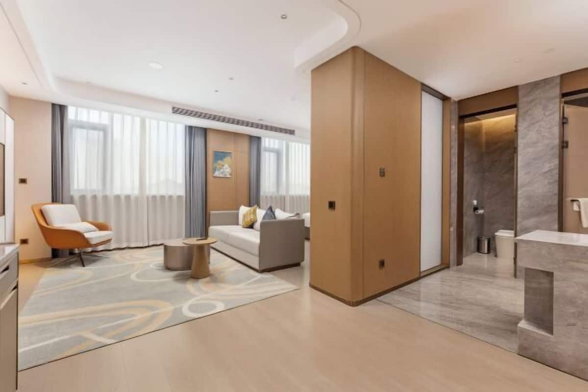 Ein modernes Hotelzimmer mit einer Couch, einem Sessel, einem kleinen Tisch, einem Badezimmer mit Spiegel und einem Kleiderschrank.