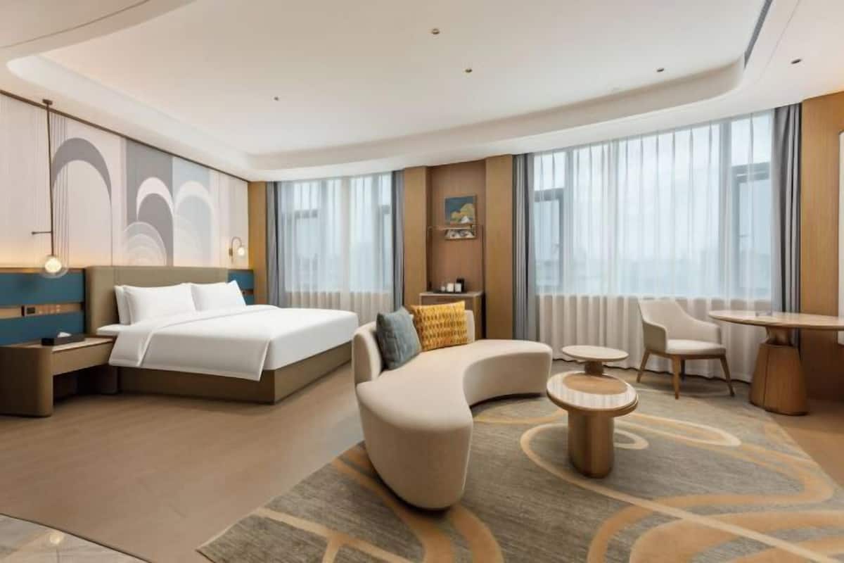 Ein modernes Hotelzimmer mit einem großen Bett, einer gebogenen Couch, einem runden Tisch und Blick auf die Stadt durch große Fenster.
