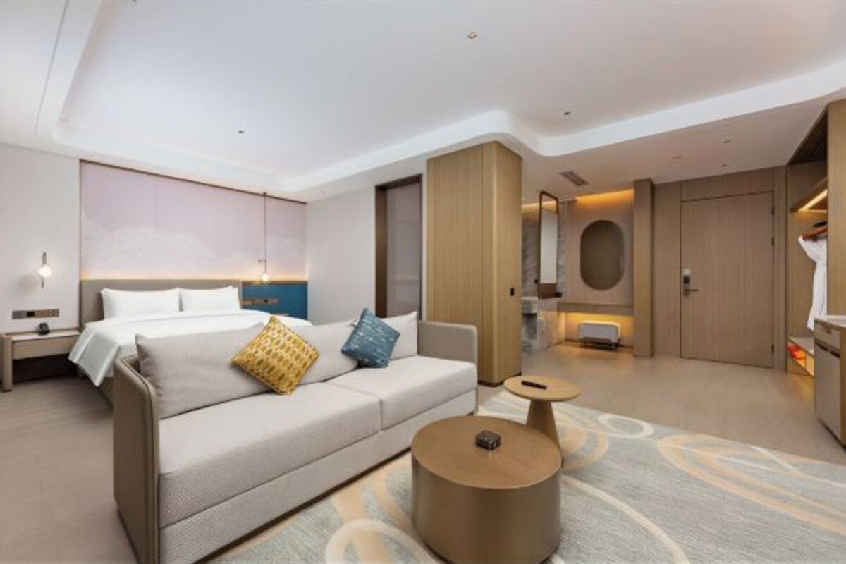 Quarto de hotel moderno com cama, sofá, mesa de centro redonda e um espelho grande.