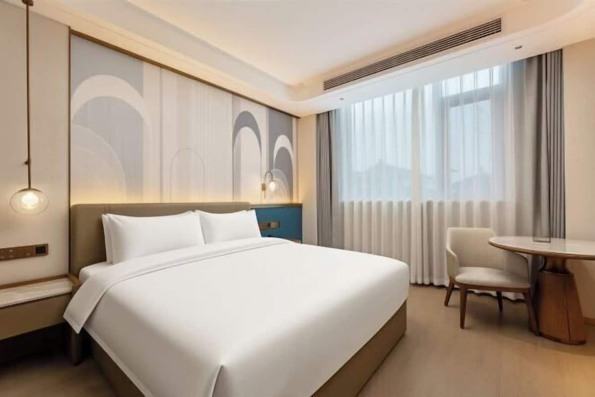 Ein Hotelzimmer mit einem großen Bett, einem Nachttisch, einem Sessel, einem runden Tisch und einem Fenster mit Vorhängen.
