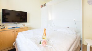 A hotel room with a bed, a television on a wooden cabinet, and a bedside table with a bottle and glasses. - Apartment No.1 - Baltic Sea Crab - Ferienhof Kluvetasch (Neustadt in Holstein)