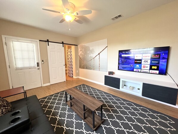 A living room with a ceiling fan, a mounted TV displaying various channels, a patterned rug, and a sliding door leading to another room.