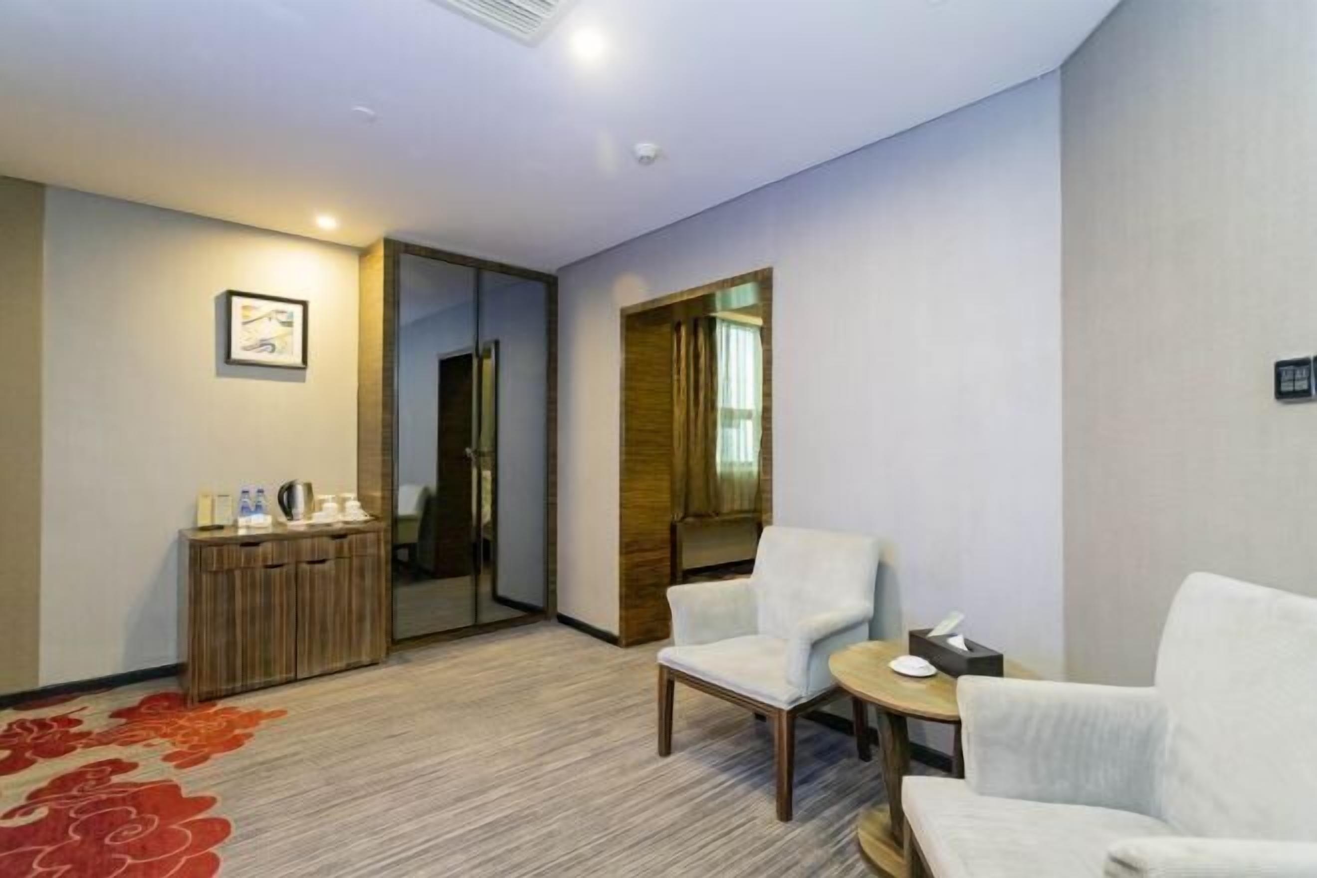 A hotel room with a wooden cabinet, two white armchairs, a small round table, and a red carpet.