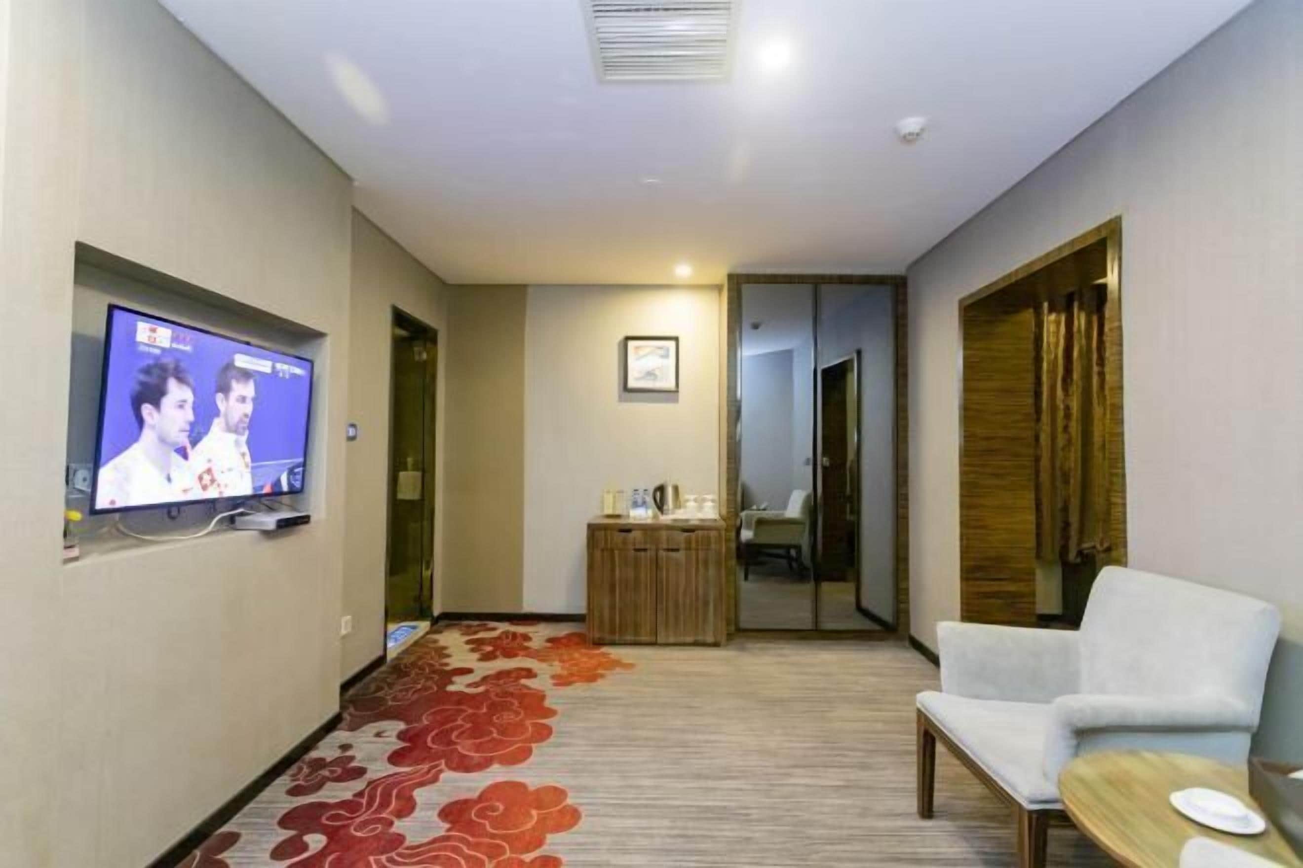 A hotel room with a television on the wall, a seating area with a chair and table, and a carpeted floor.
