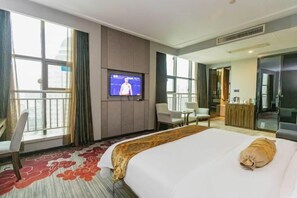 A hotel room with a large bed, a television mounted on the wall, a desk with a chair, and a view of the city through the windows. - Beiliu International Hotel (Beiliu)