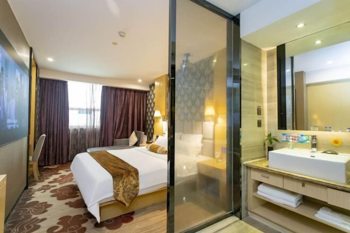 A hotel room with a large bed, a glass partition, a bathroom with a white sink, and a large mirror.
