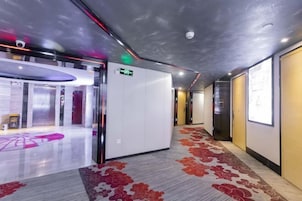 Un pasillo de hotel con alfombra estampada, una zona de recepción con separador de vidrio y un ascensor moderno.